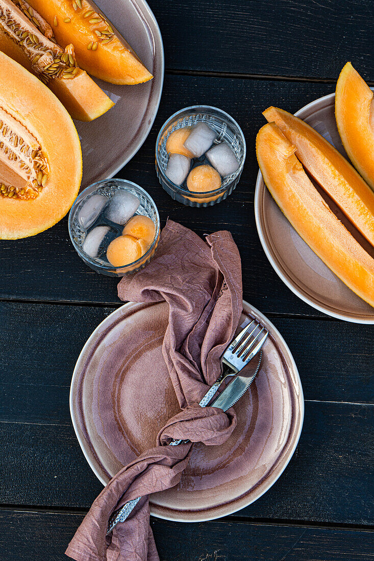 Top view of sliced organic fresh orange musk melon fruit and melon spheres in transparent glasses with ice cubes while served on table with napkin fork and knife in daylight