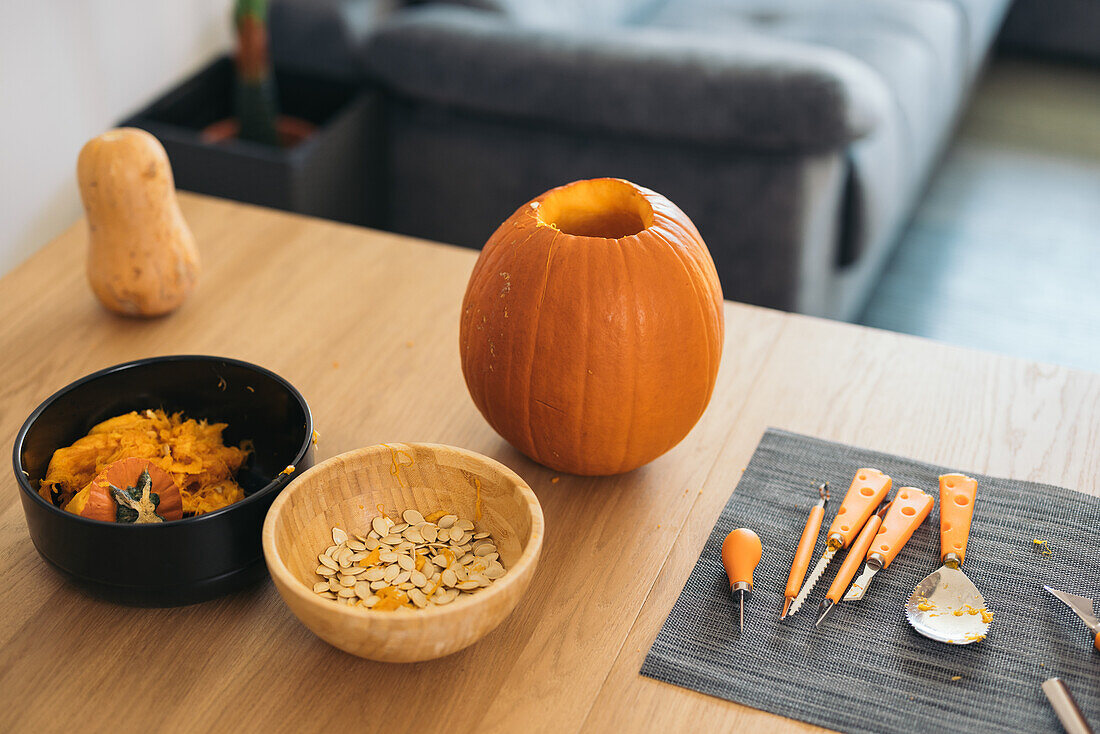 High angle of Orange pumpkin for Halloween occasion and bowls with seeds and pulp placed on wooden table against blurred sofa at home