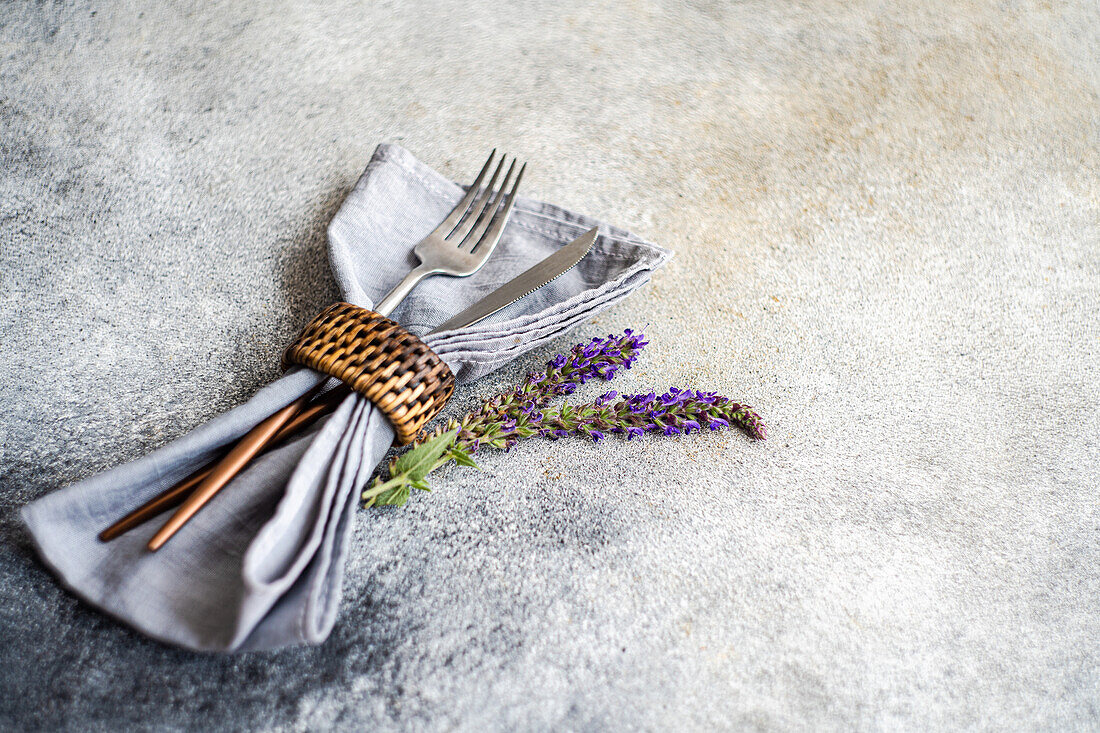 Hoher Winkel eines gedeckten Tisches mit Silberbesteck, Serviette und Lavendelblüten vor grauem, unscharfem Hintergrund