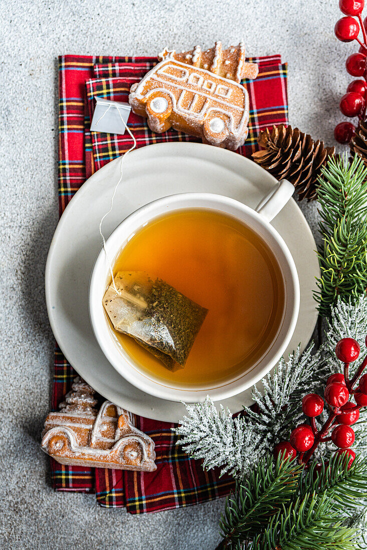 Draufsicht auf eine weihnachtliche Tasse Tee auf einer roten Serviette neben Stechpalme, Keksen und Tannenzapfen auf einem grauen Tisch