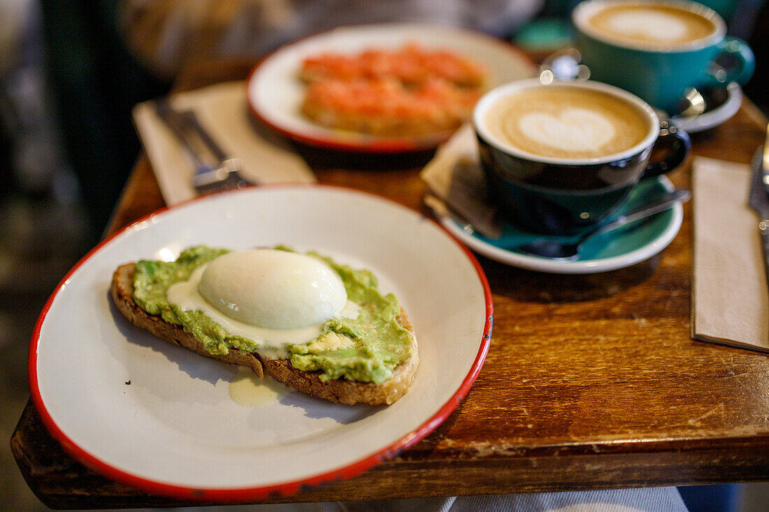 Delicious toast with poached egg and smashed avocado on ceramic plate near cups of cappuccino in cafe