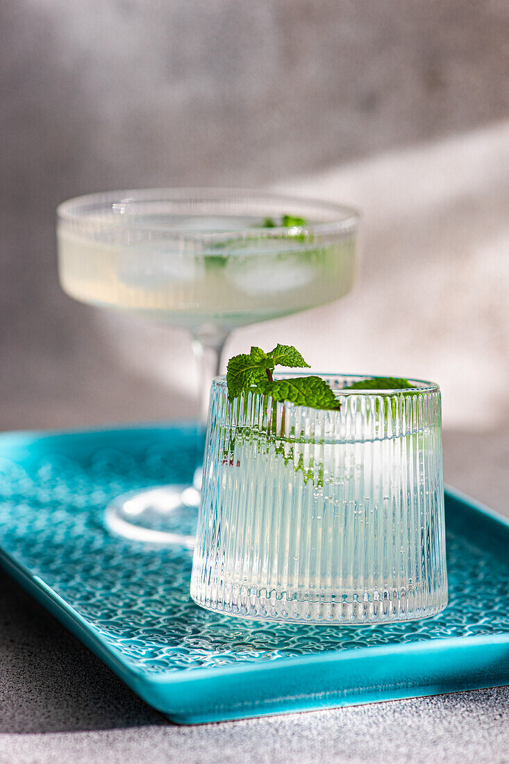 Two elegant cocktails on a blue tray with a textured background, one garnished with mint