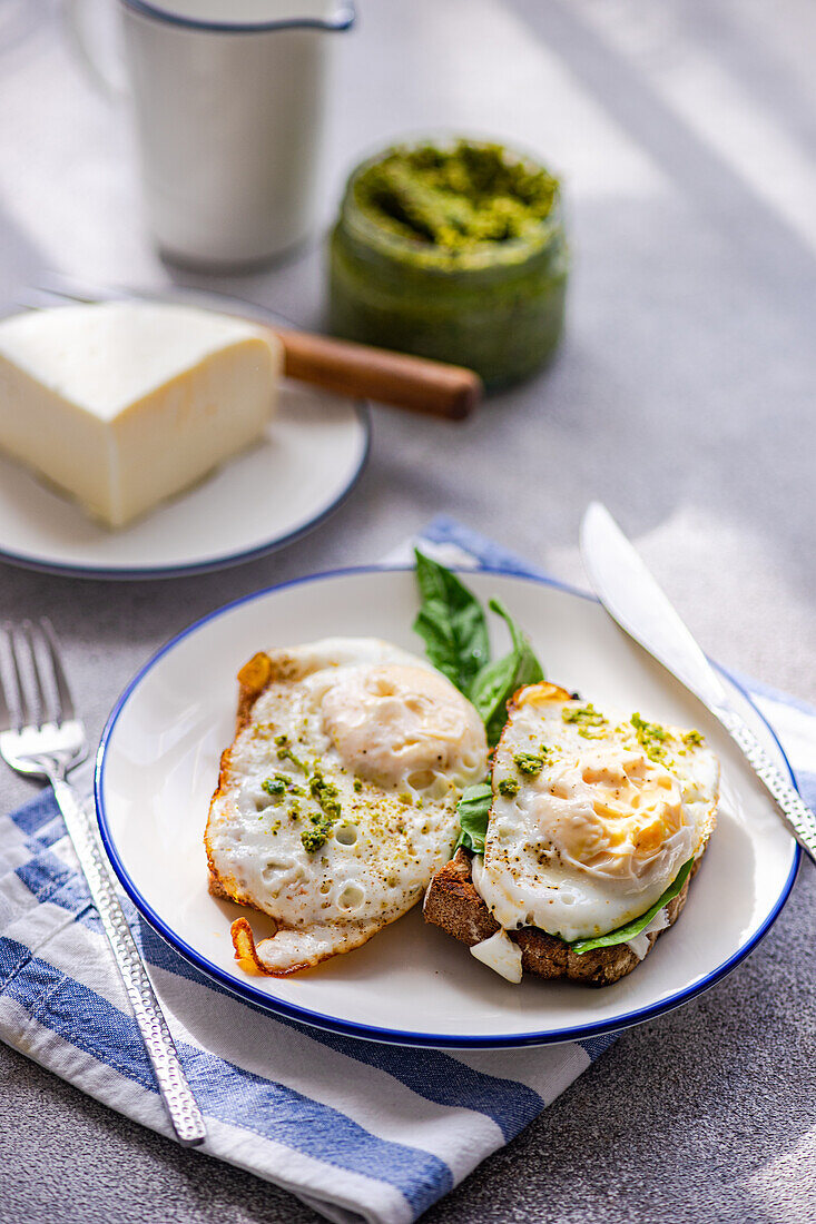 Blick von oben auf einen Teller mit gesundem Toast mit Spiegelei-Pesto-Sauce, grünen Kräutern und Käse auf einem grauen Tisch beim Frühstück