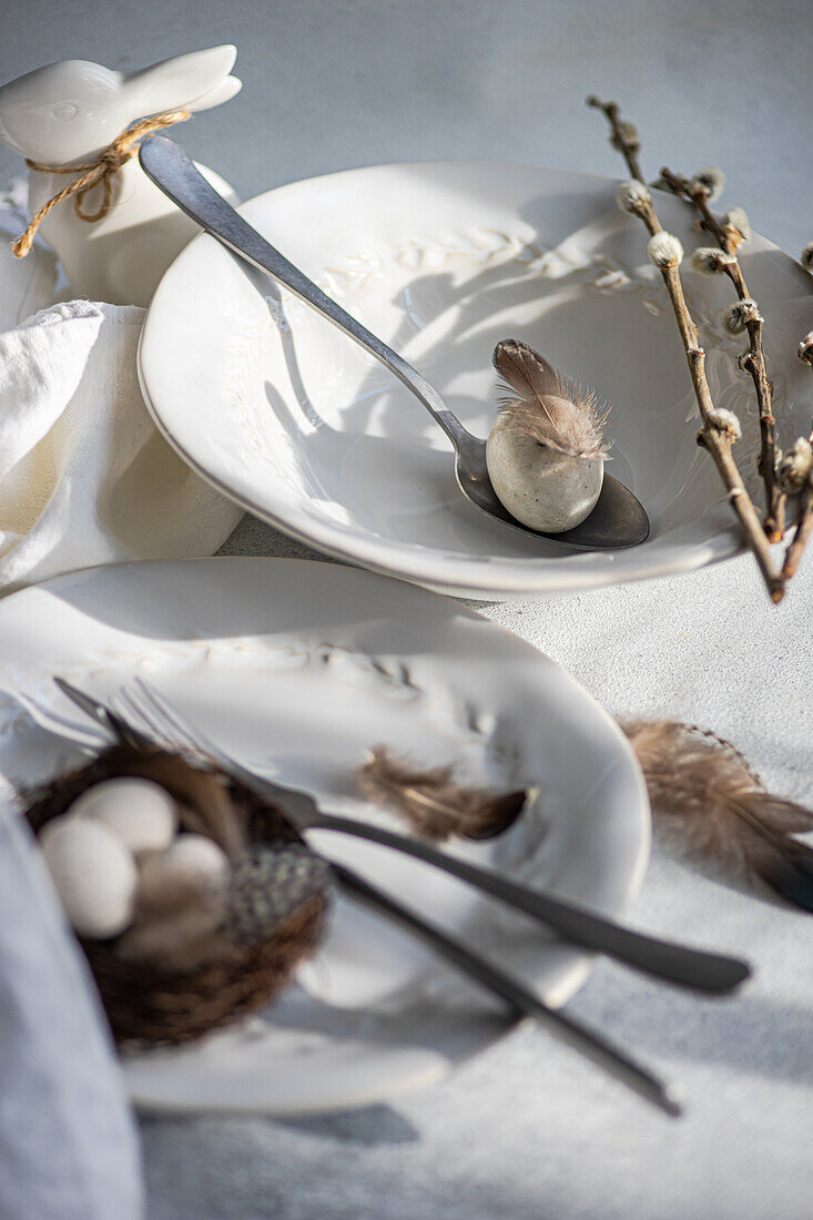 Nahaufnahme eines eleganten Osterarrangements mit makellosen weißen Tellern mit zartem Prägemuster und einem gewebten Nest mit gesprenkelten Eiern auf einem Teller, begleitet von Gabel und Messer, Federn und Zweigen