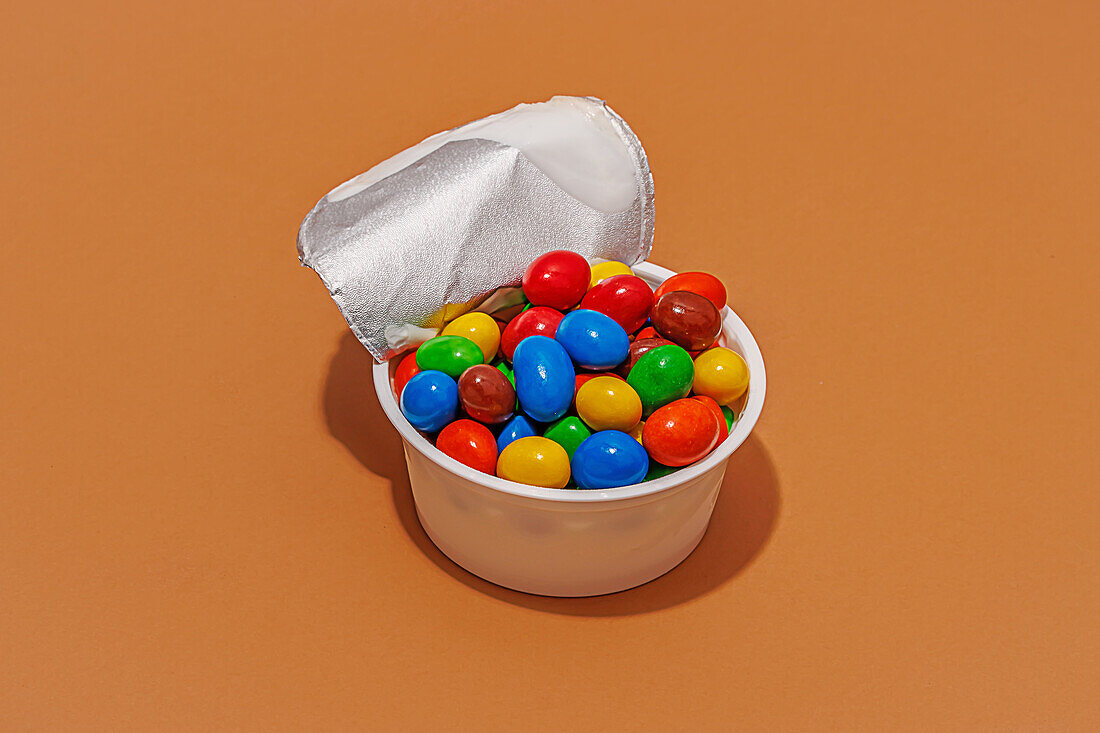 High angle of opened yoghurt container filled with colored candies against orange background