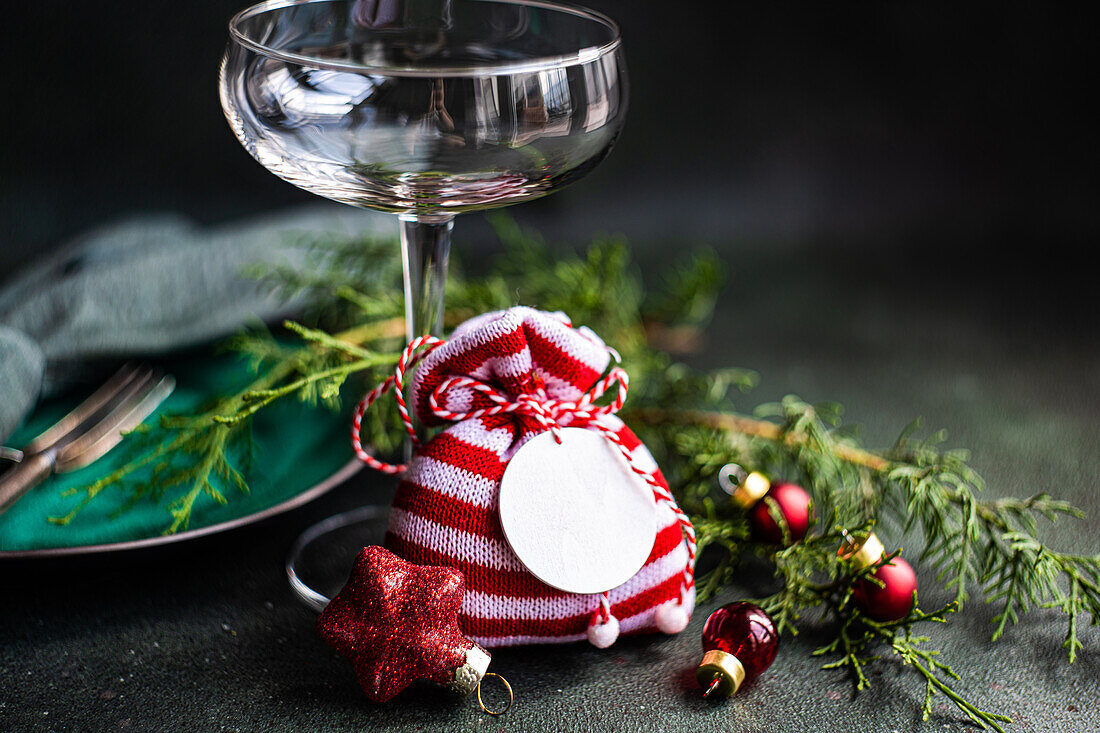 Glass with small red and white bag placed near fir sprigs on dark surface in Christmas time