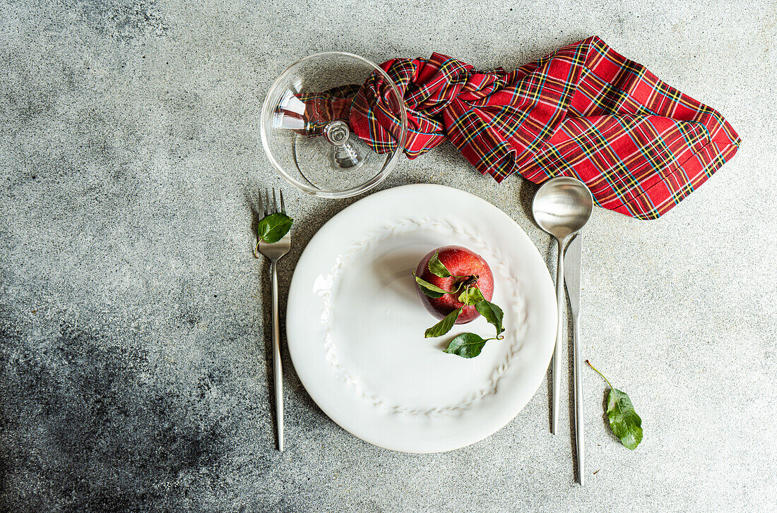 Top view of ripe red apple in plate placed between knife,spoon and fork with cloth and empty glass on gray surface