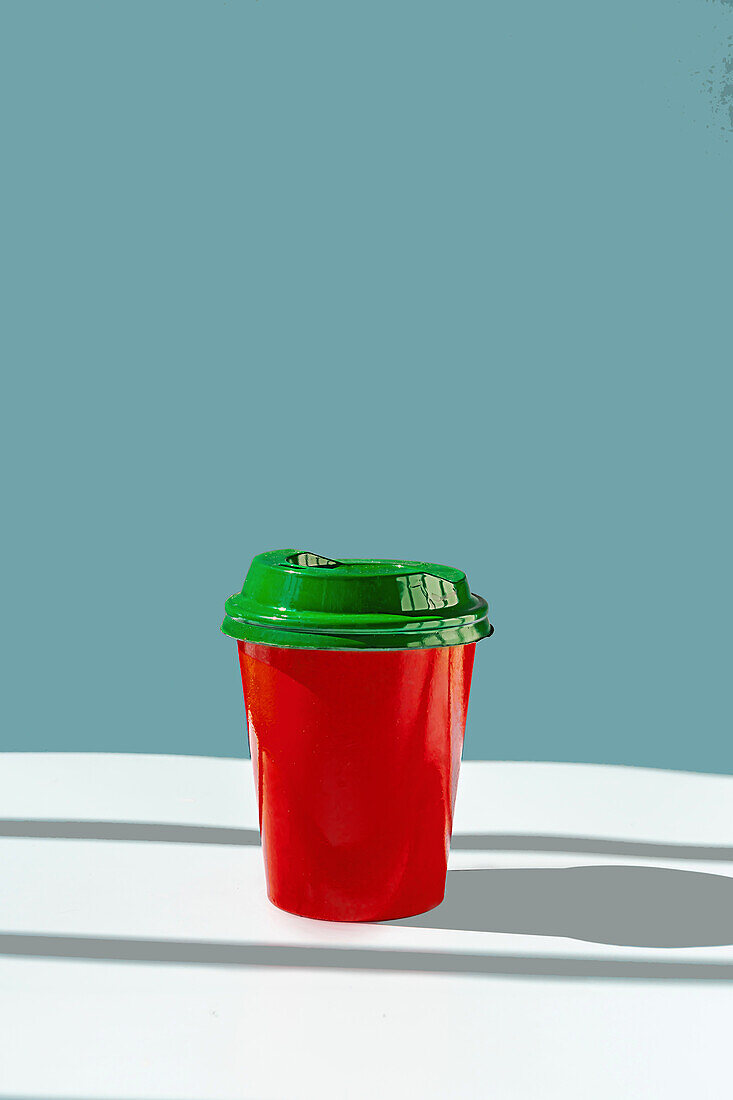 Minimalistischer roter Plastikbecher mit Kaffee zum Mitnehmen und grünem Deckel vor blauem Hintergrund