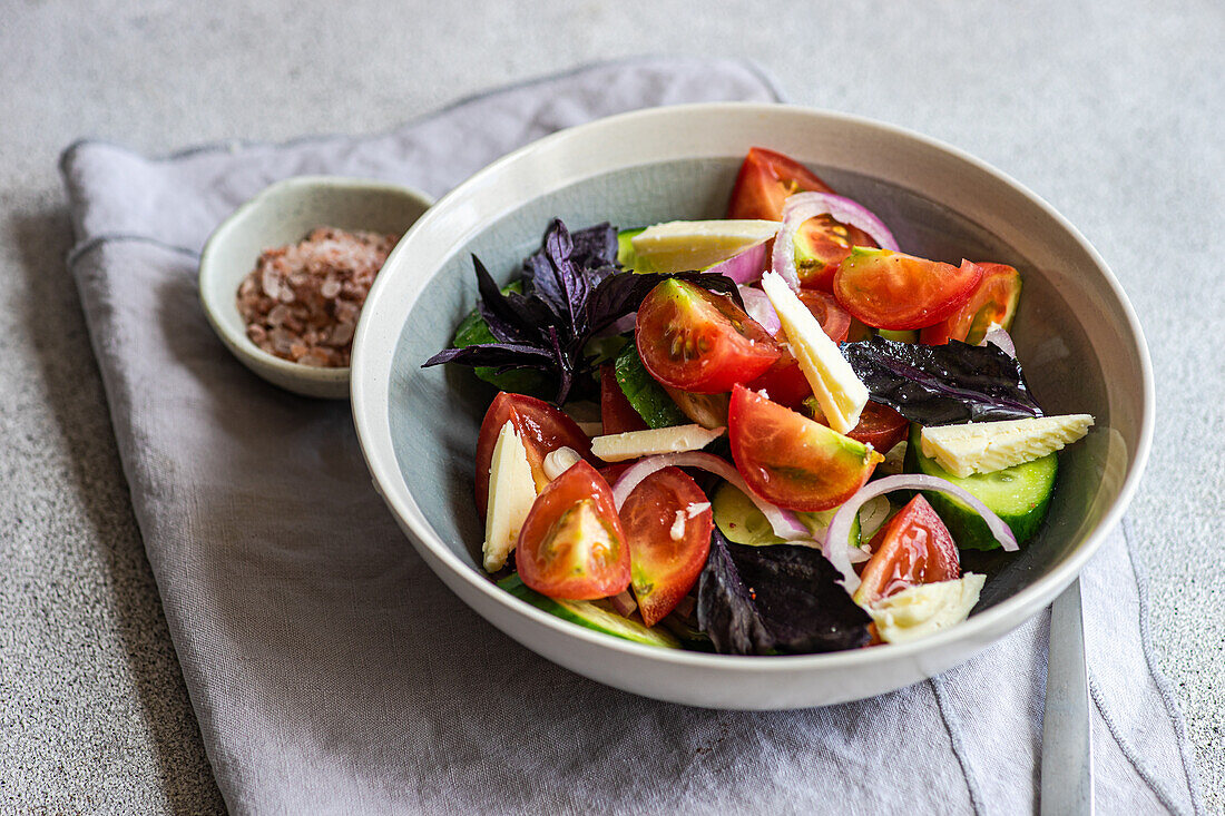 High angle of healthy vegetable salad with basil leaves and cheese slices in bowl on gray surface with napkin and salt