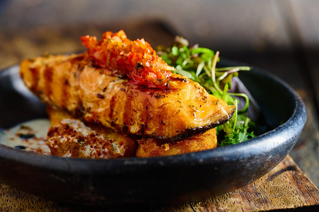 Front view of appetizing grilled salmon steak garnished with sauce and herbs served on black bowl placed on wooden cutting board