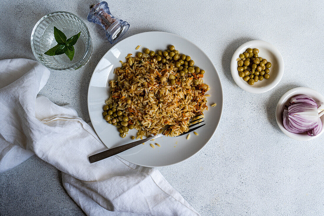 Draufsicht auf gekochten Reis mit Zwiebel, Karotte, Paprika, grünen Erbsen und Gewürzen auf einem Teller mit Gabel neben Zwiebelscheiben und einem Glas Wasser mit Minze vor weißem Hintergrund