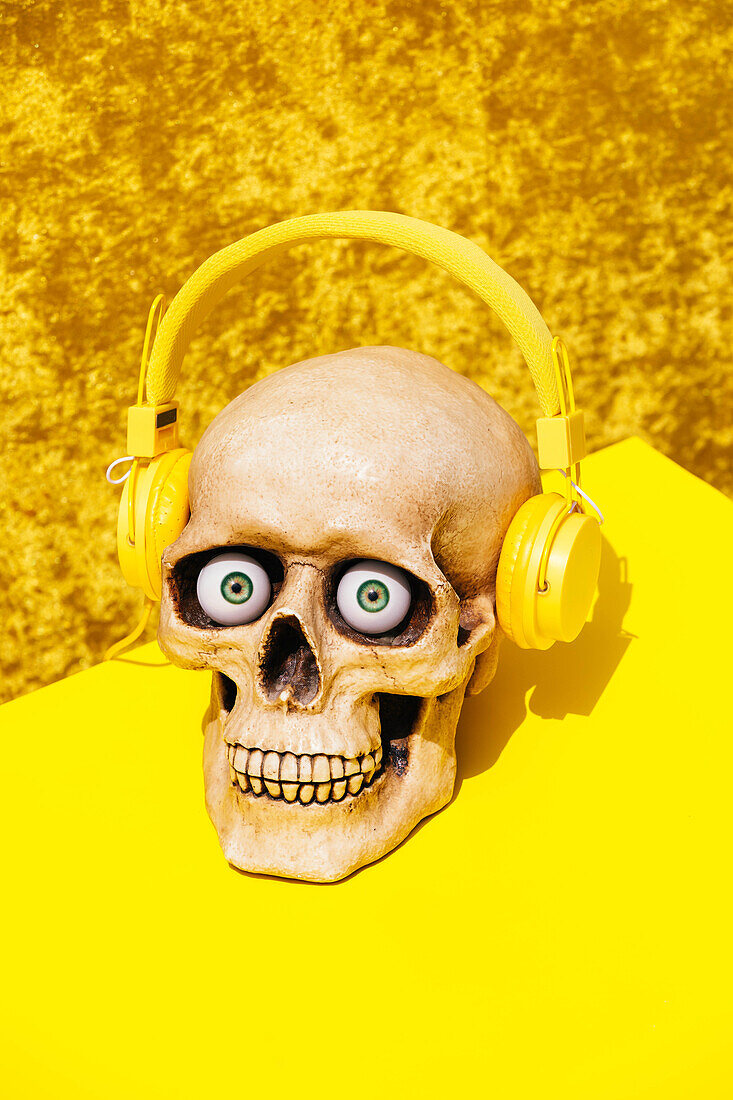 Gruseliger menschlicher Schädel mit drahtlosen Kopfhörern, der mit Augäpfeln in die Kamera schaut, von oben auf leuchtend gelbem Hintergrund