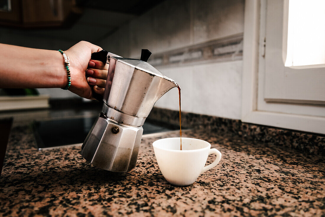 Anonyme Hand mit türkisfarbenem Armband gießt frisch gebrühten Kaffee aus einer Espressomaschine auf dem Herd in eine weiße Tasse vor einem modernen Küchenhintergrund