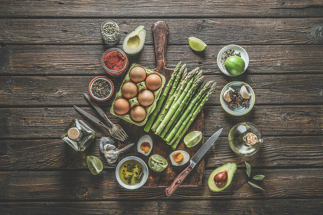 Various healthy cooking ingredients: green asparagus bunch, eggs, spices, avocado, lime, olive oil and kitchen utensils: forks, knifes and cutting board on rustic wooden table. Top view.