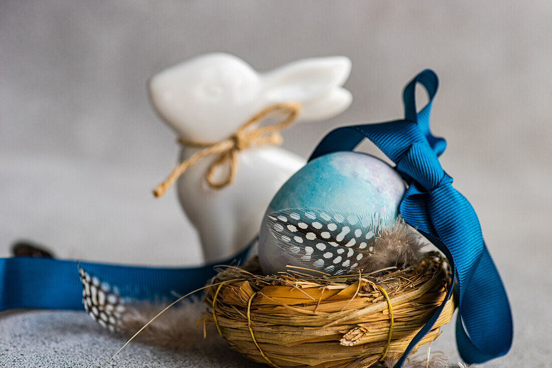 Eine Nahaufnahme einer Osterkomposition mit einem blau gefärbten, mit einem Band geschmückten Ei in einem Nest, einer Perlhuhnfeder und einem Keramikhasen auf einem strukturierten Hintergrund