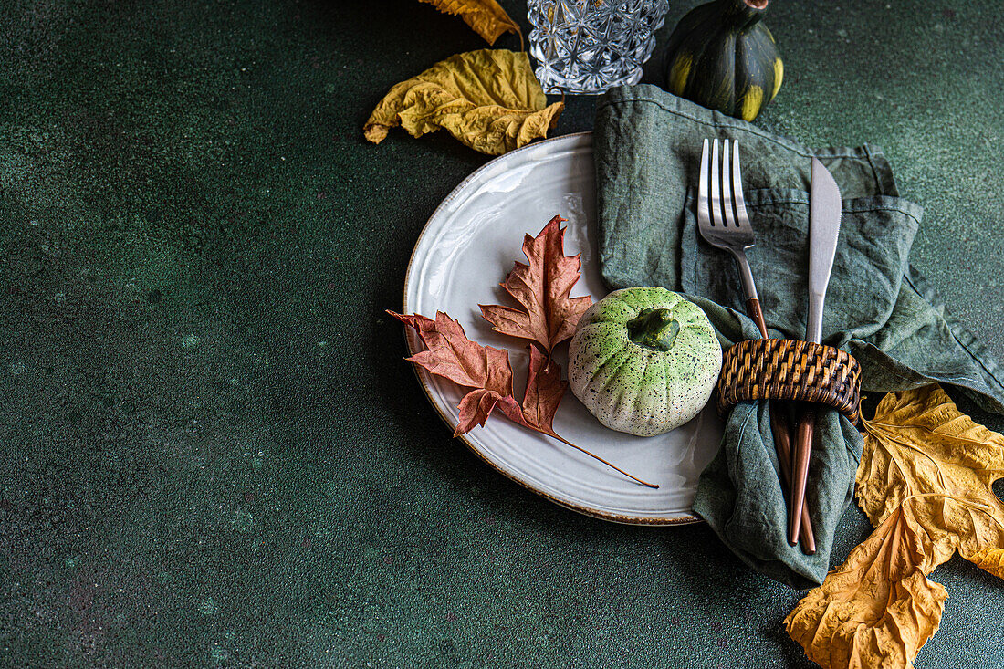 High angle of autumnal table setting with napkin, knife and fork place on plate near colorful leaves, smalls pumpkins and empty glass against dark surface