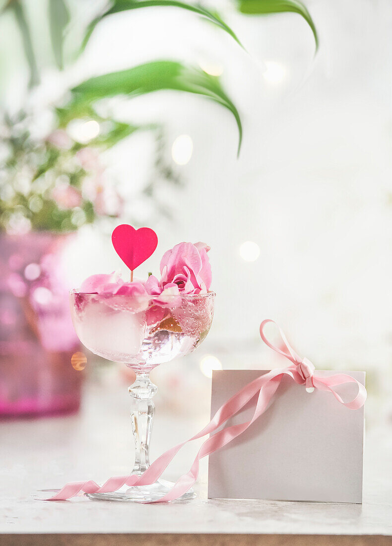 Champagne glass with pink rose petals, heart and blank white gift card with pink ribbon on white table at floral bokeh background. Celebrating valentines day. Front view