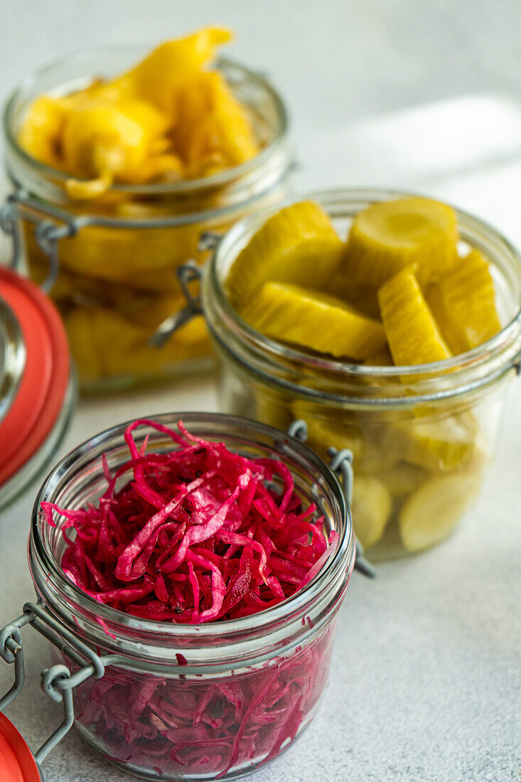 A vibrant display of fermented vegetables, featuring cabbage with beetroot, hot spicy peppers, and white cucumber pickles in glass jars.
