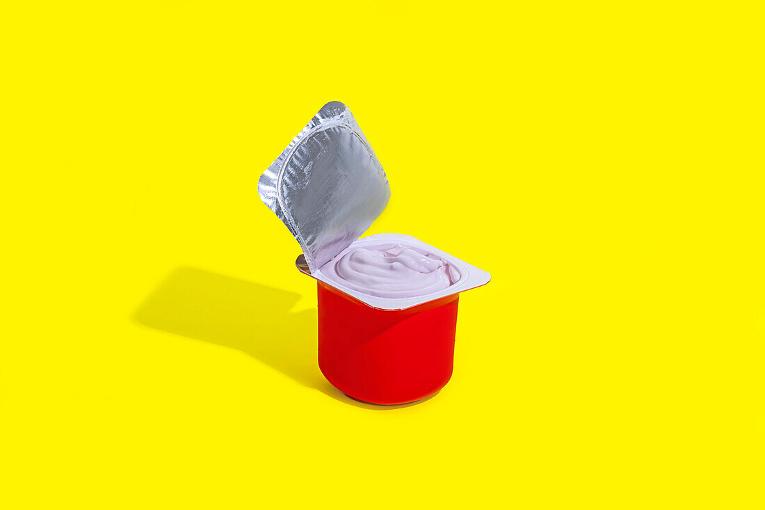 Ein einsamer roter Becher mit Erdbeerjoghurt, dessen Foliendeckel teilweise abgezogen ist, steht vor einem leuchtend gelben Hintergrund