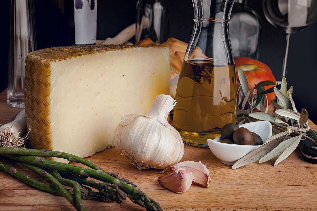 Handwerklich hergestellter Käse neben Knoblauch, Spargel, Oliven und Olivenöl, bereit für die Zubereitung einer anspruchsvollen Mahlzeit