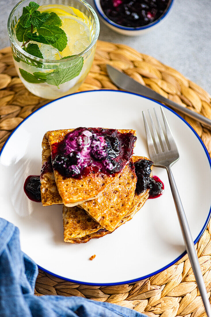 Stack of homemade crepes topped with sweet blueberry jam and a dollop of sour cream and lemon water drink served on a white plate with a blue rim.