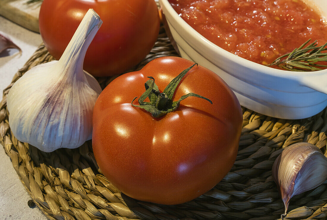 Close up of bowl with tomato spread and fresh tomatoes placed on table near unpeeled garlic