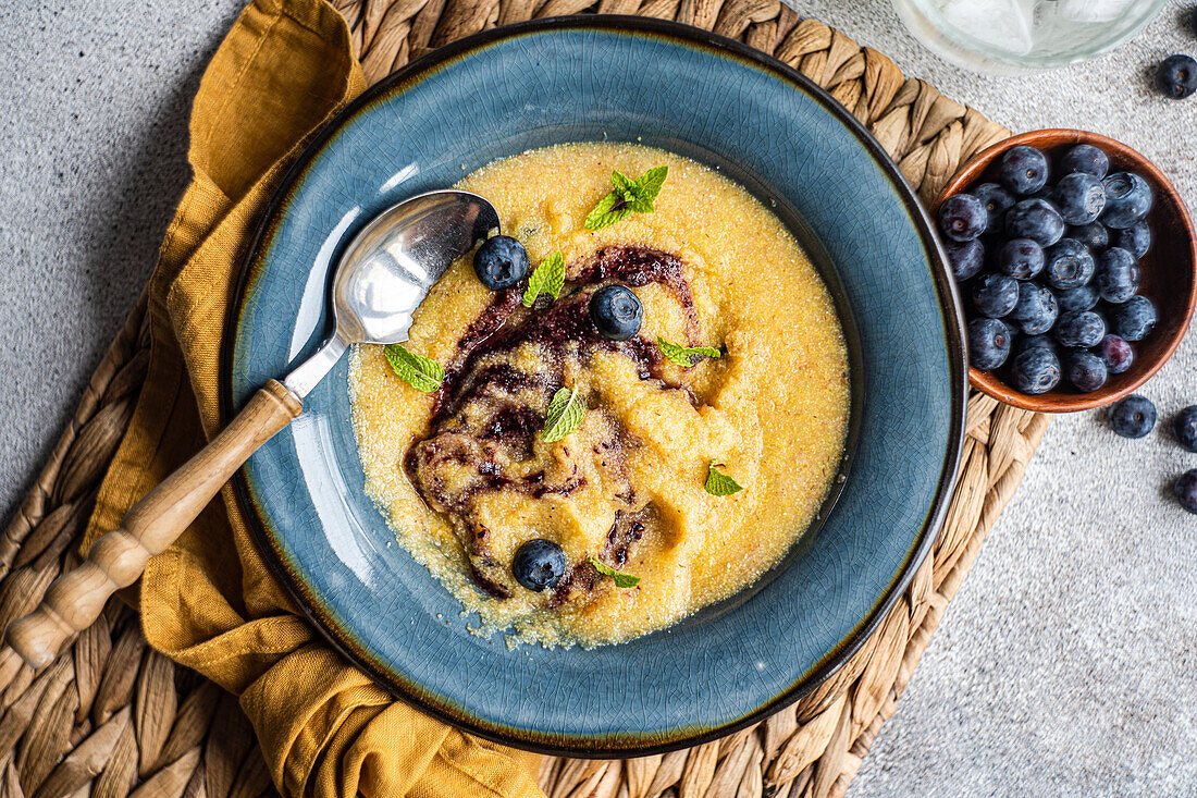 Top view of polenta porridge with mint and jam near fresh blueberries in bowl on table