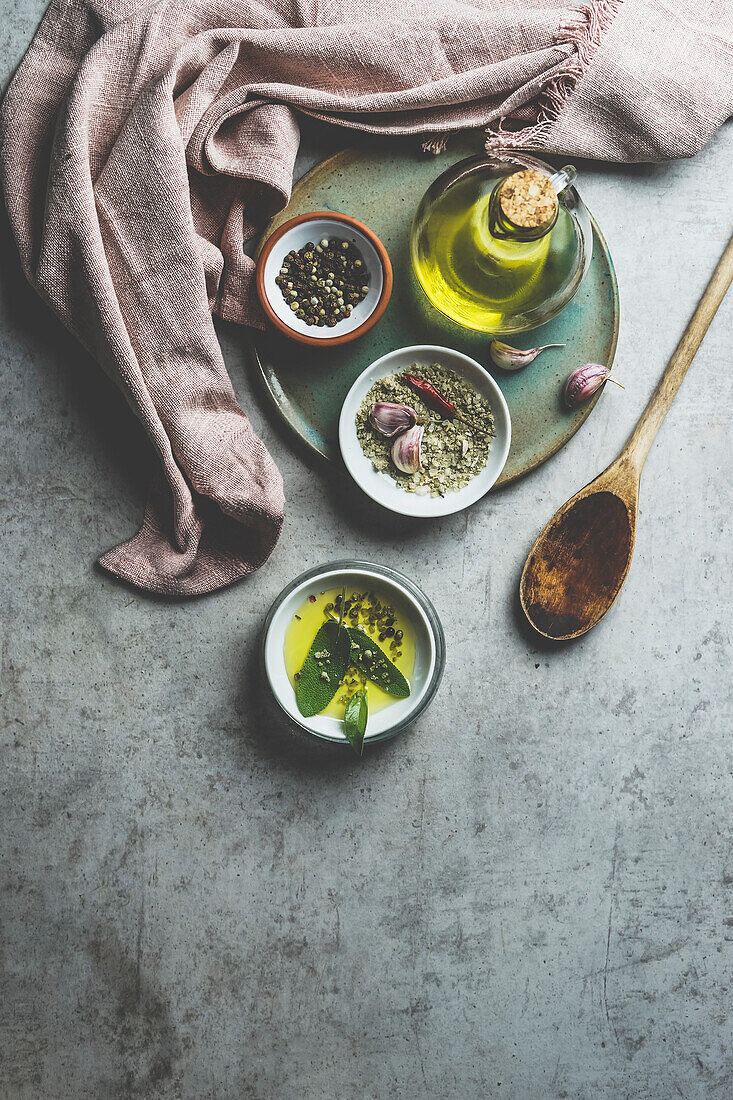 Healthy cooking ingredients on grey concrete kitchen table: olive oil, sage, garlic, pepper, salt, wooden cooking spoon, dish cloth. Cooking flavorful food at home. Top view.