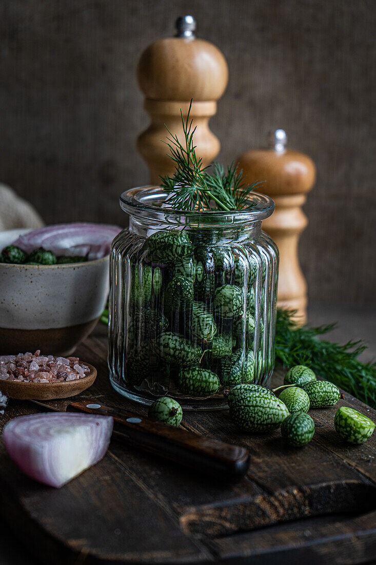 Zutaten für die Zubereitung von fermentierten Gurken in einem Glas auf einem Holztablett neben einer Serviette und Salz- und Pfefferstreuern vor dunklem Hintergrund