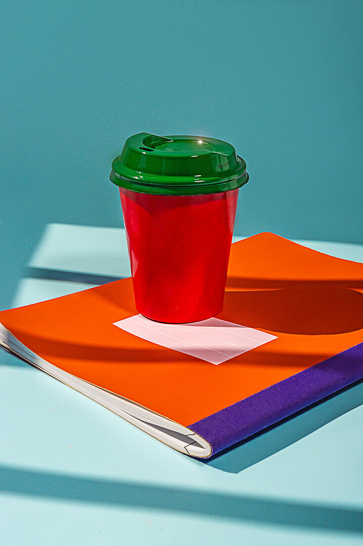 Minimalistischer roter Plastikbecher mit Kaffee und grünem Deckel auf dem Tisch über einem Notebook vor blauem Hintergrund