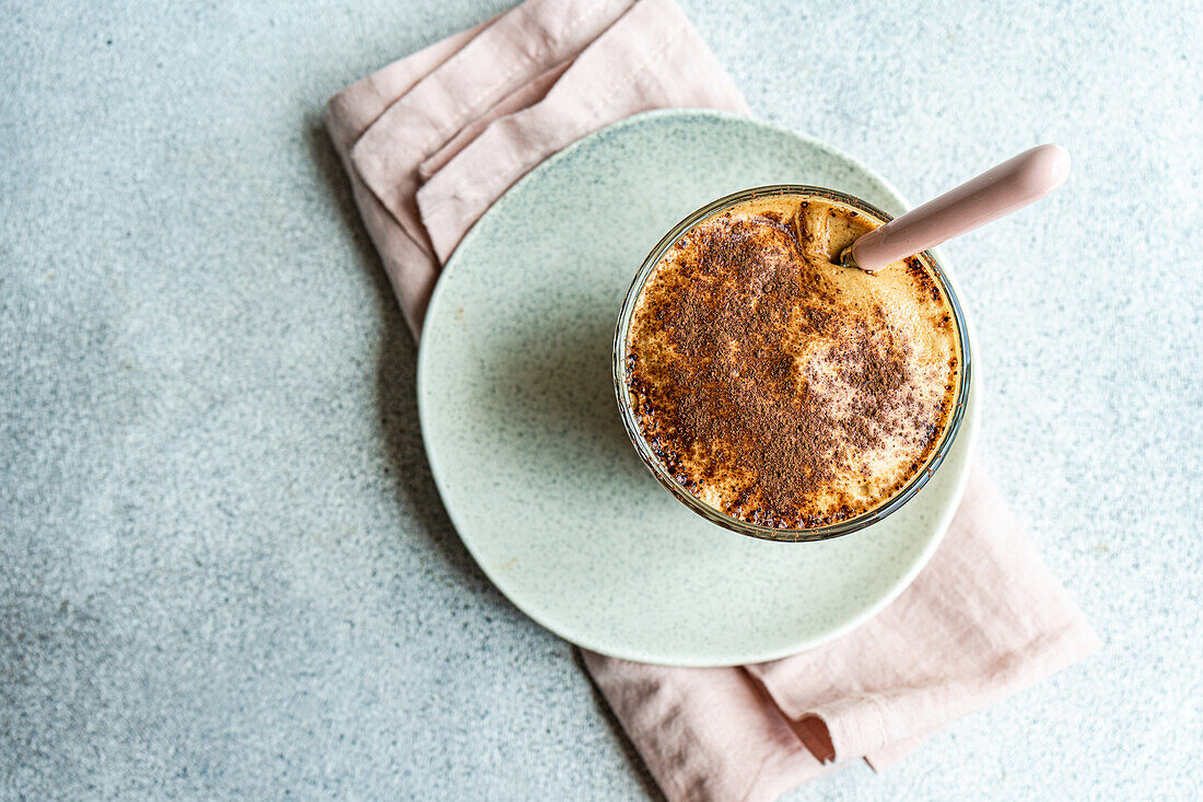 Top view of glass cup with coffee cream dessert with chocolate powder and ice served on plate with spoon on napkin for refreshing summer meal against blurred background