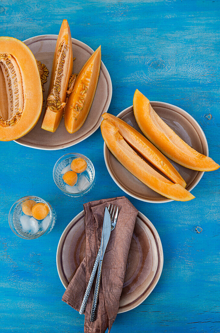 Draufsicht auf in Scheiben geschnittene frische Bio-Orangen-Moschus-Melone und Melonenkugeln in transparenten Gläsern mit Eiswürfeln, serviert auf einem Tisch mit Serviette, Gabel und Messer bei Tageslicht