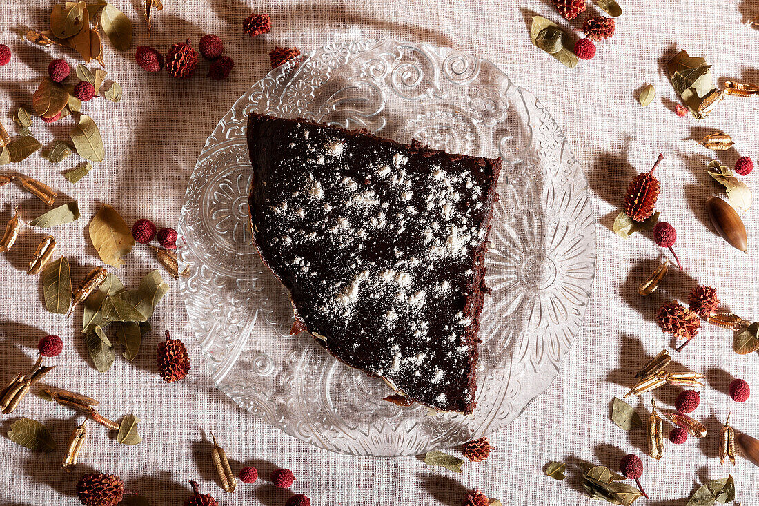 Draufsicht auf ein köstliches Stück Schokoladenkuchen, bestäubt mit Puderzucker, auf einem verzierten Teller, umgeben von getrockneten Blütenblättern