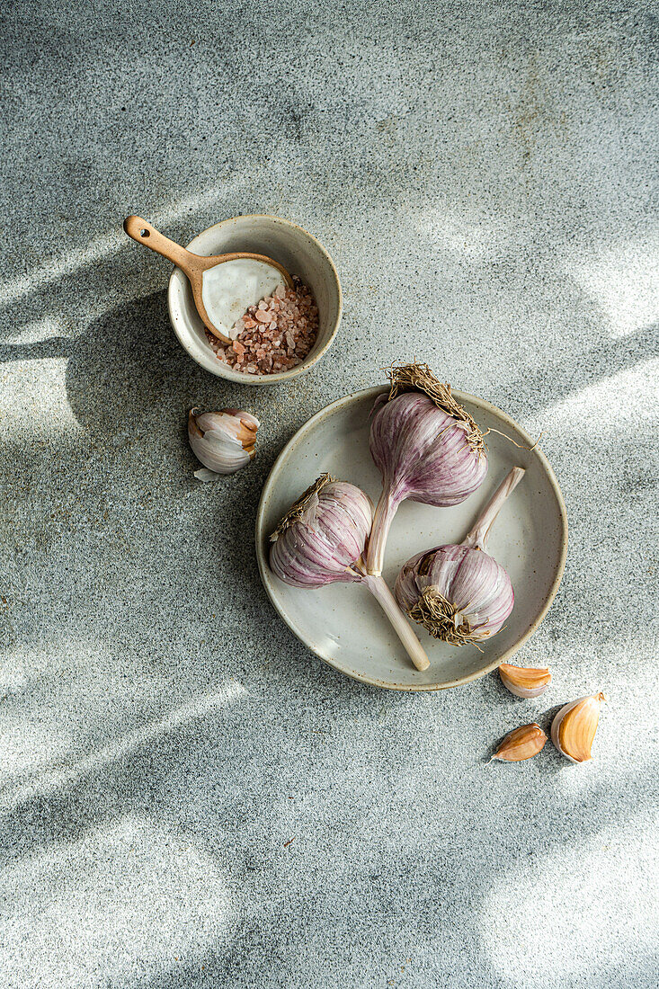 From above of raw garlic bulbs with a bowl of coarse salt on a textured grey surface illuminated by soft sunlight casting shadows