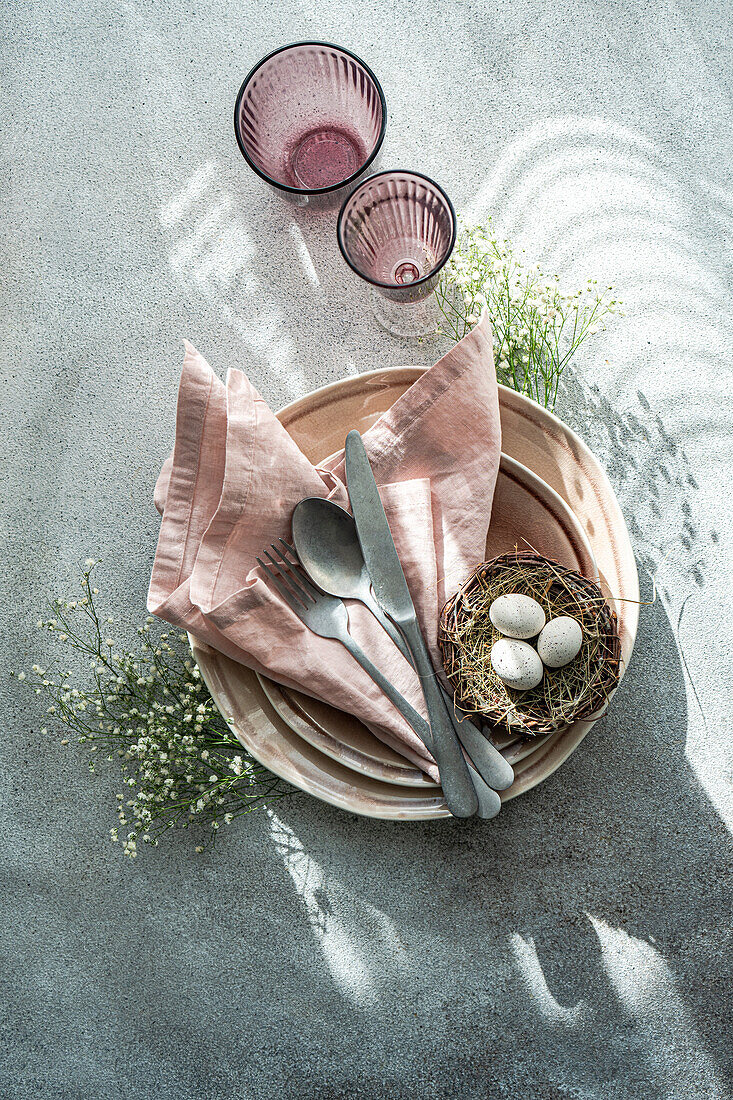Top view elegant and simple table setting celebrates Easter with a bird's nest containing speckled eggs, surrounded by delicate china and soft pink linen.