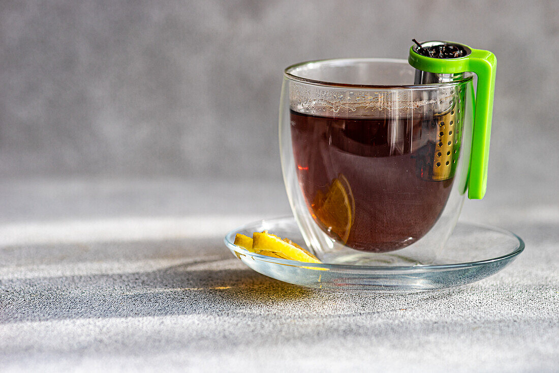 A transparent cup of hot tea with a slice of lemon on a saucer and a green tea infuser hanging on the side