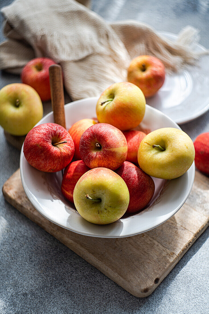 From above of collection of ripe, colorful apples presented on a rustic wooden board, with a white plate, fork, and draped fabric creating a serene kitchen setting