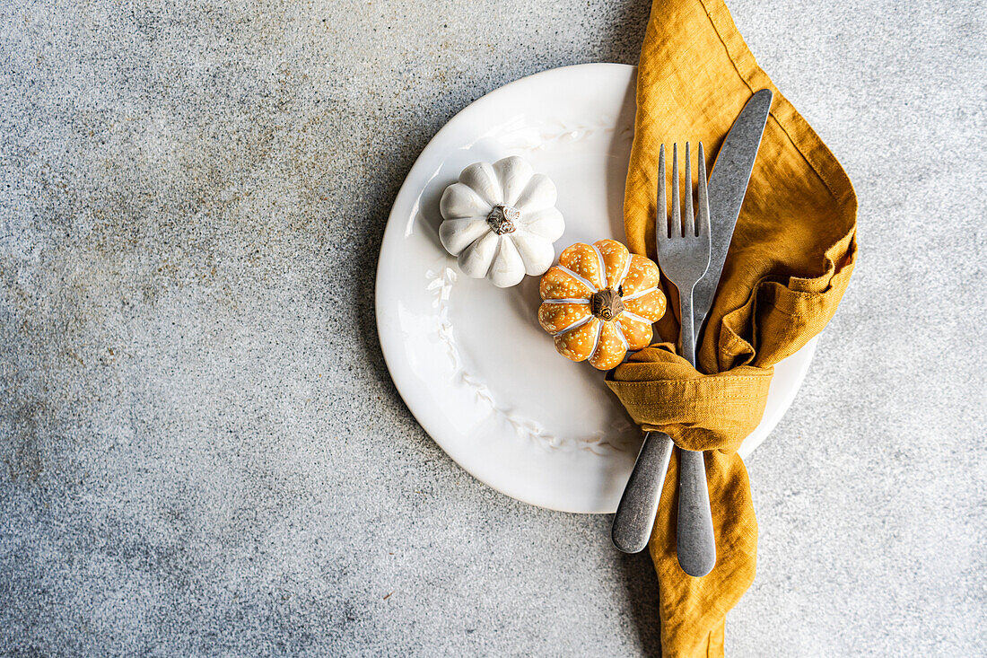 Top view of white ceramic plate with decorative pumpkins with silver fork and knife, wrapped in a mustard-colored napkin, placed on a textured gray concrete surface