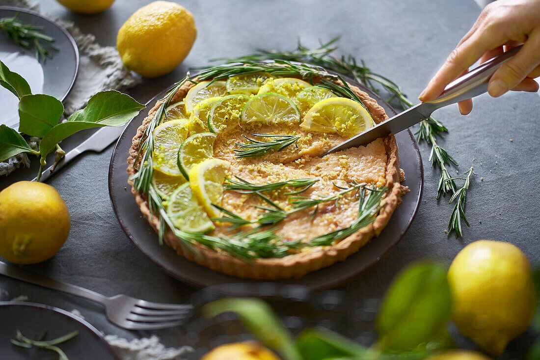 From above anonymous person cutting round lemon pie with decorated with lemon slices and rosemary sprigs on table in the kitchen