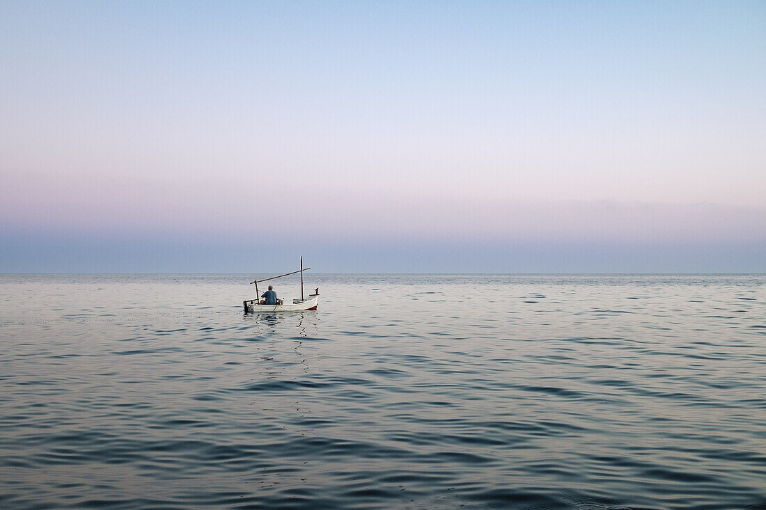 Anonymer Fischer auf einem einsamen schwimmenden Boot auf dem plätschernden Meerwasser von Soller auf Mallorca beim Fischen vor dem wolkenlosen blauen Himmel bei Sonnenuntergang im hellen Abendlicht