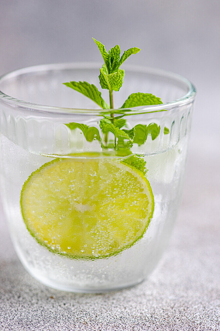 Glass of summer lemonade with tonic and fresh lime and mint leaves placed on gray surface during summer season at home