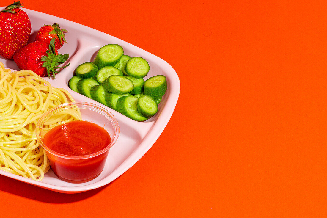 From above composition of spaghetti with tomato sauce cucumber slices and yummy fresh strawberries served for school lunch on orange top table