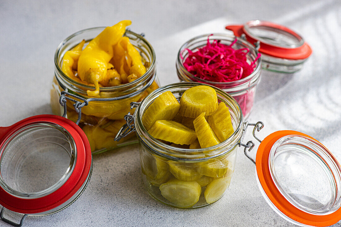 Three glass jars filled with vibrant fermented vegetables, including yellow hot peppers, red cabbage with beetroot, and green pickled cucumbers, displayed on a light surface.