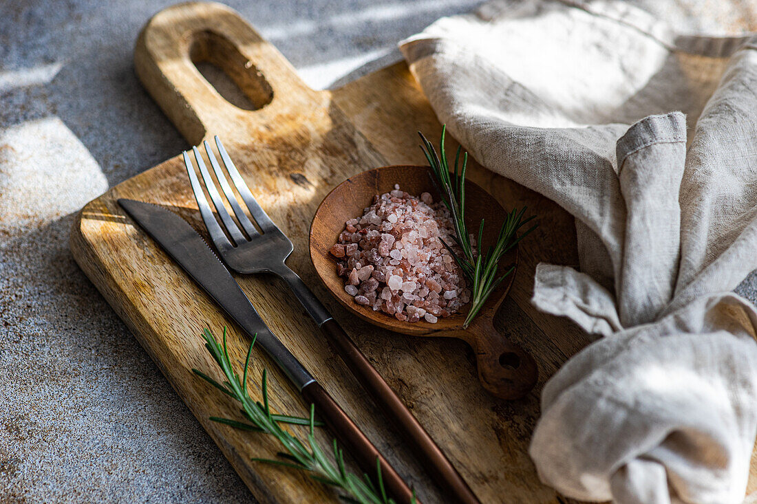 From above rustic kitchen setup on wooden cutting board with a fork, knife, a sprinkle of pink Himalayan salt, fresh rosemary sprigs, and a linen napkin, laid out on a textured concrete background