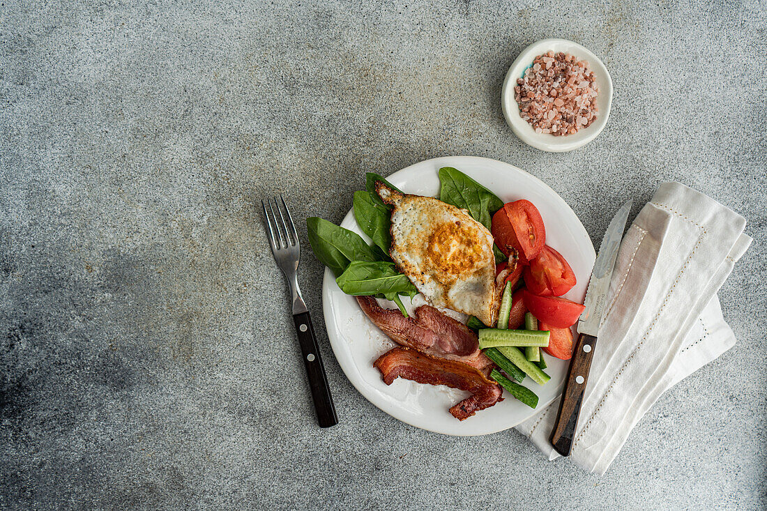 Draufsicht auf ein ausgewogenes Frühstücksarrangement auf einem weißen Teller mit Spiegelei, knusprigem Speck, frischem Blattspinat, Gurkenscheiben und keilförmigen Tomaten, serviert mit Besteck auf einer grau strukturierten Oberfläche