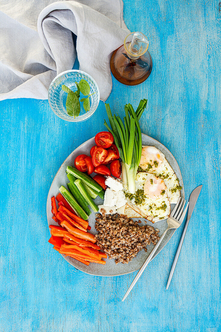 A colorful breakfast plate featuring sliced cherry tomatoes, mini cucumbers, green onions, and red bell pepper strips, paired with crumbled feta cheese, boiled buckwheat, and fried eggs drizzled with pesto sauce.