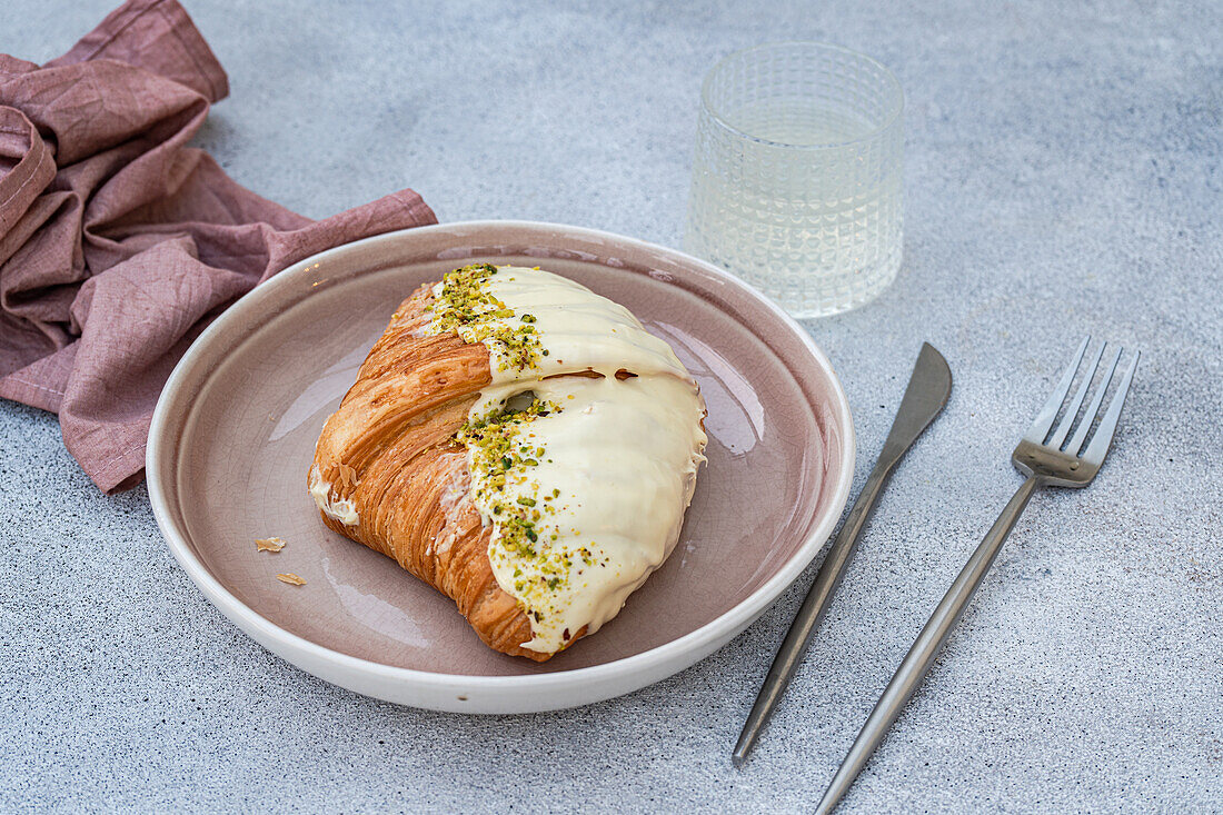 Goldenes Croissant von oben, teilweise mit weißem Zuckerguss und Pistazienstücken bedeckt, auf einem runden beigen Teller neben Messer und Gabel, auf grauem Hintergrund mit einer gefalteten braunen Serviette