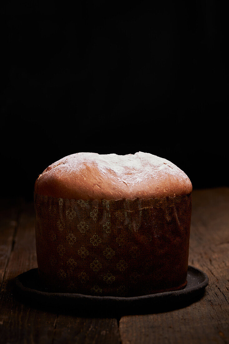 A freshly baked panettone sits on a rustic wooden table, highlighted against a dark background, creating an intimate and inviting scene