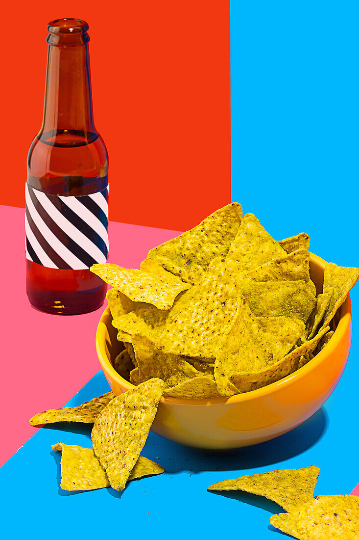 Ein lebhaftes Stillleben mit einer Schüssel knuspriger Nacho-Chips neben einer dunklen Flasche vor einem geteilten Hintergrund aus Rot und Blau