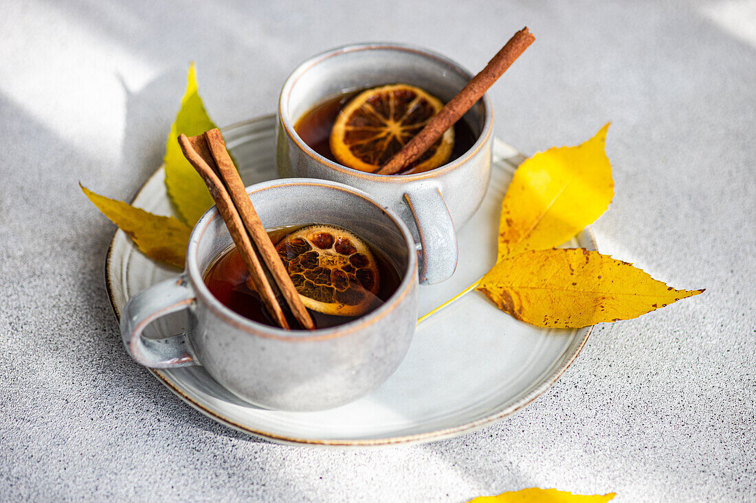 Two cups of aromatic spiced tea with cinnamon sticks, anise, and dried orange slices surrounded by radiant yellow autumn leaves on a textured gray background